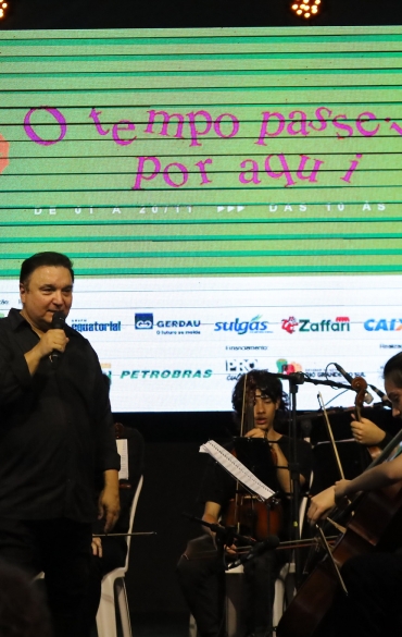 A imagem retrata um palco durante um evento musical. No centro, um homem de cabelos curtos e escuros, vestido com uma camisa preta, segura um microfone e aparenta estar falando ao público. Atrás dele, há músicos sentados, incluindo violinistas e violoncelistas, com partituras posicionadas em suportes. À direita, uma musicista toca violoncelo, e outros integrantes da orquestra são visíveis ao fundo.

No painel luminoso ao fundo, há o texto "O tempo passeia por aqui" em destaque, sobre um fundo listrado em tons de verde. Abaixo, são exibidos logotipos de patrocinadores e apoiadores do evento, como Gerdau, Sulgás, Zaffari, Caixa, Petrobras e outros. A iluminação no palco é suave, com refletores no topo. O ambiente está repleto de espectadores, visíveis na parte inferior da imagem.