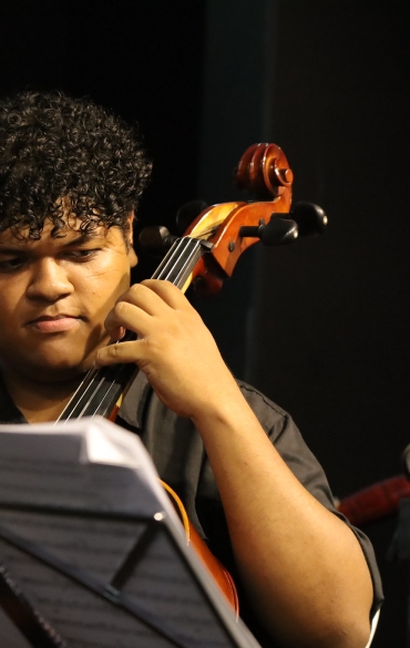 
A imagem mostra um músico tocando violoncelo. Ele está em foco e parece concentrado enquanto olha para as partituras à sua frente. Ele tem cabelos curtos e cacheados, veste uma camisa preta e segura o arco com a mão direita enquanto a esquerda está no braço do instrumento. O fundo está desfocado, mas aparenta ser um ambiente interno com iluminação amarela suave. Um microfone em suporte é visível ao lado direito, parcialmente iluminado. A partitura no suporte à frente do músico está levemente inclinada.