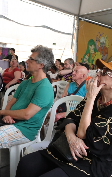 A imagem mostra uma plateia composta por pessoas de diferentes idades e estilos, sentadas em cadeiras brancas de plástico, assistindo a um evento. O espaço é coberto por uma tenda, proporcionando sombra aos espectadores. Em primeiro plano, uma mulher com boné laranja e óculos escuros, vestindo um traje preto com detalhes dourados, aparece pensativa, com a mão apoiada no rosto. Ao lado dela, um homem de camisa verde e shorts xadrez parece atento ao que está sendo apresentado.

Ao fundo, pode-se observar mais espectadores, com alguns rostos voltados para o palco. A decoração da área inclui painéis coloridos com ilustrações e mensagens, como "Não existe potente sem cultura". A estrutura conta ainda com arquibancadas, onde mais pessoas estão acomodadas. O ambiente transmite uma atmosfera de envolvimento cultural e contemplação coletiva.