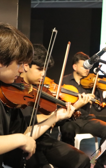 A imagem mostra um grupo de músicos tocando violino em um ambiente fechado. O foco principal está em um jovem com cabelo castanho liso, vestindo uma camisa preta de manga curta, enquanto toca seu violino com concentração. Atrás dele, outros músicos, também vestidos de preto, estão posicionados com seus violinos. Há um microfone montado em um pedestal no canto direito da imagem, direcionado para o grupo. O ambiente parece ser um palco ou espaço de apresentação, com um fundo escuro e iluminação voltada para os músicos. Na parte de trás, há uma tela com textos e gráficos parcialmente visíveis. O clima é formal, sugerindo uma apresentação musical organizada.