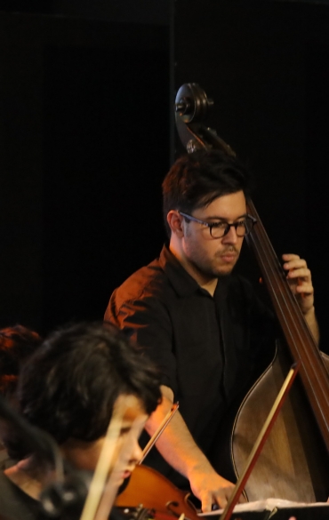 A imagem retrata uma apresentação musical com foco em um músico tocando contrabaixo. Ele é um homem jovem, usando óculos e vestido com uma camisa preta, concentrado no instrumento. Ao seu redor, outros músicos da orquestra são parcialmente visíveis, tocando diferentes instrumentos, como violinos e violoncelos. O fundo é escuro, indicando um ambiente fechado e provavelmente um palco. Há partituras sobre os suportes, sugerindo um ensaio ou uma performance formal. A iluminação destaca os músicos contra o fundo sombreado, criando um clima íntimo e focado na música.