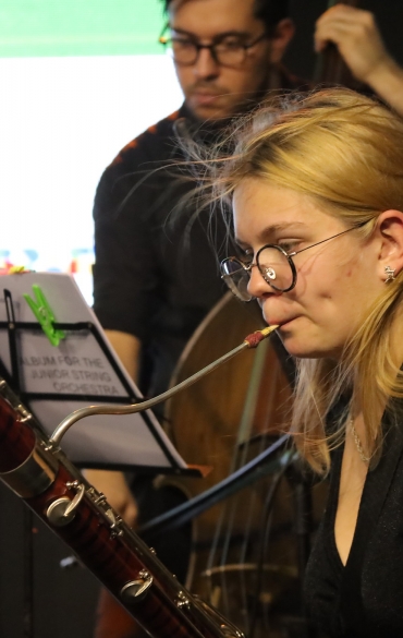 
A imagem mostra uma jovem musicista tocando fagote durante uma apresentação ou ensaio. Ela tem cabelos loiros, usa óculos redondos e está vestida com uma blusa preta. Sua expressão é focada enquanto toca o instrumento. Ao fundo, é possível ver outro músico tocando contrabaixo, parcialmente desfocado, e um suporte com partituras, que inclui uma prendedora verde. Há uma tela iluminada ao fundo, com logotipos parcialmente visíveis, sugerindo um evento formal. A composição destaca o fagote em primeiro plano, com detalhes de madeira polida e metais, e transmite um clima de dedicação musical.