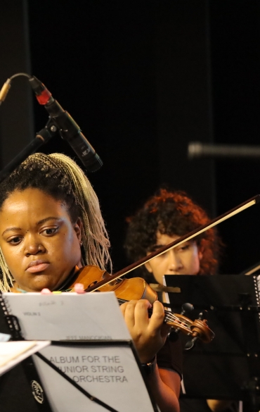 A imagem mostra um grupo de músicos tocando violinos. Em destaque, há uma mulher com tranças loiras e expressão concentrada segurando um violino, lendo uma partitura. Ao fundo, estão outros músicos também concentrados, incluindo um homem à esquerda e duas pessoas à direita, todos segurando violinos. Eles parecem estar em um ensaio ou apresentação, com microfones e suportes de partituras visíveis. O ambiente é fechado, com iluminação suave e fundo escuro.