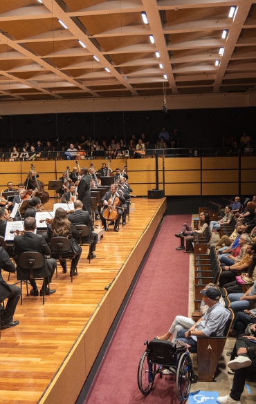 A imagem mostra uma apresentação de uma orquestra sinfônica em um auditório. No palco de madeira, músicos tocam diversos instrumentos como violinos, violoncelos, contrabaixos e outros, enquanto o maestro, em pé, rege a orquestra. Os músicos estão alinhados em semicírculo, vestindo trajes formais. À frente do palco, há uma plateia lotada, com pessoas sentadas em cadeiras de fileiras organizadas. No canto inferior direito, observa-se uma pessoa em uma cadeira de rodas, indicando acessibilidade no local. O teto do auditório é composto por um design geométrico com luminárias embutidas, e as paredes são revestidas com painéis de madeira. A iluminação destaca o palco e cria um ambiente acolhedor e elegante.