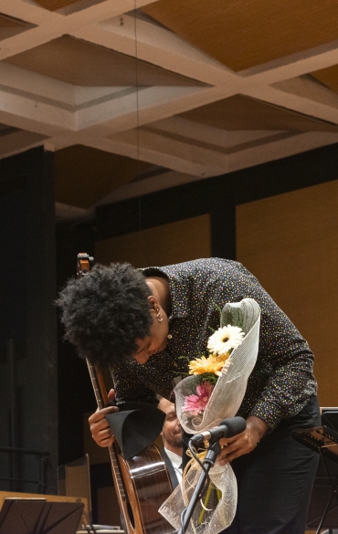 Uma pessoa inclina-se em agradecimento ao público, segurando um violão clássico e um buquê de flores coloridas envolto em papel decorativo. Ela está sorrindo discretamente, demonstrando gratidão, enquanto outros músicos ao fundo também sorriem, observando o gesto. A pessoa veste uma camisa preta com estampas coloridas e está em pé ao lado de uma cadeira e um microfone. O ambiente é de uma sala de concerto, com detalhes em madeira e uma atmosfera calorosa e de celebração musical.