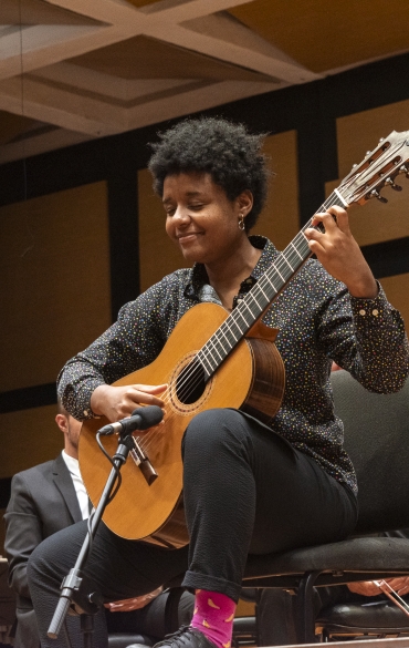 Uma pessoa tocando violão clássico em um palco de concerto. Ela está sentada em uma cadeira, segurando o instrumento com expressão de concentração e leve sorriso. Usa uma camisa preta com estampas coloridas, calça preta, e sapatos escuros, destacando-se suas meias cor-de-rosa com detalhes amarelos. Ao fundo, é possível ver músicos e seus instrumentos, além de uma estrutura de madeira e metal, típica de salas de concerto. Um microfone está posicionado próximo ao violão. O ambiente transmite uma atmosfera de performance ao vivo e apreciação musical.