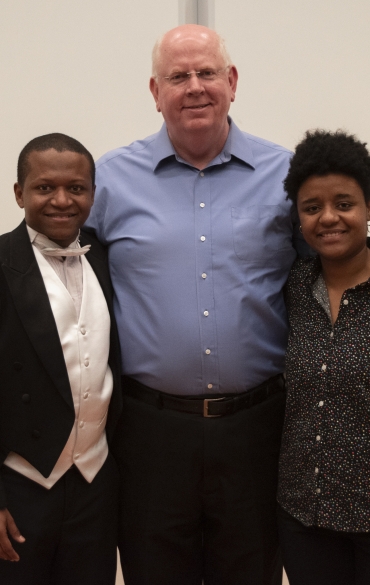 A imagem apresenta três pessoas posando lado a lado. À esquerda, um homem negro vestindo um fraque preto, camisa branca e gravata borboleta branca. Ele está sorrindo. Ao centro, um homem branco de pele clara, com cabelos curtos e calvos na parte superior, usando uma camisa social azul e calça preta. Ele sorri e abraça as pessoas ao seu lado. À direita, uma mulher negra de cabelo curto e crespo, vestindo uma camisa de manga longa preta com estampa de pequenos pontos coloridos e uma calça preta. Ela também sorri, e as mãos de ambos os homens estão apoiadas em seus ombros. O fundo é neutro, com uma parede clara.