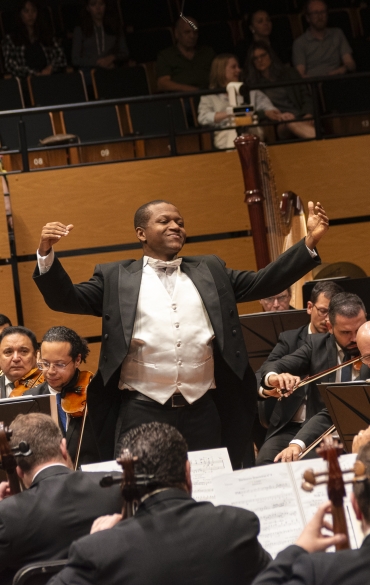 Um maestro conduz uma orquestra sinfônica em uma sala de concerto. Ele está de pé no centro do palco, vestindo um traje formal preto com colete branco, com os braços levantados em um gesto expressivo. À frente, a orquestra é composta por músicos tocando instrumentos de corda, como violinos, violas e violoncelos. Ao fundo, é possível ver harpas e outras seções orquestrais. A plateia, sentada nas arquibancadas superiores, observa atentamente a apresentação. O ambiente é elegante, com painéis de madeira na parede e iluminação quente.