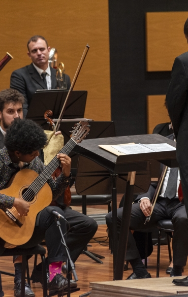 Um músico tocando violão clássico em um concerto, sentado em frente a uma partitura. Ele está em destaque, com expressão concentrada e vestindo camisa preta com estampas coloridas, calça preta e meias rosas com detalhes amarelos. À sua frente, o maestro está de costas para a câmera, vestindo um fraque preto e conduzindo a apresentação. No fundo, os músicos da orquestra estão sentados, atentos, com seus instrumentos e partituras. O ambiente é uma sala de concerto com painéis de madeira nas paredes, criando uma atmosfera formal e dedicada à música erudita.