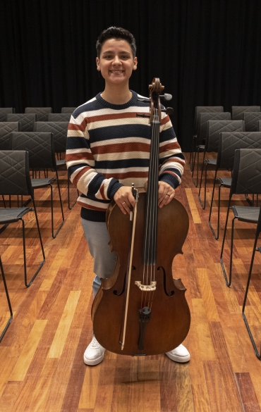 
A imagem mostra uma pessoa jovem sorrindo, em pé, segurando um violoncelo em uma sala de apresentação ou ensaio. Ela veste um suéter de listras horizontais nas cores branca, azul-marinho, laranja e bege, junto com calça jeans clara e tênis brancos. O violoncelo está posicionado à sua frente, e ela segura o arco com uma das mãos. O chão é de madeira, e ao fundo há várias cadeiras vazias com estofado escuro e um cortinado preto cobrindo a parede. A atmosfera é acolhedora e iluminada, típica de um ambiente musical.