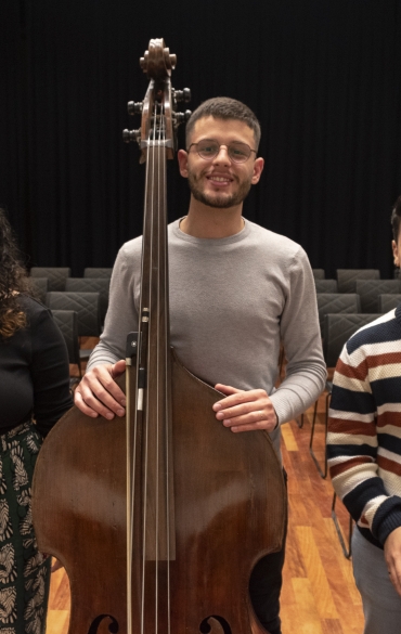 A imagem apresenta três jovens músicos posando em um auditório. À esquerda, uma jovem segura uma flauta transversal e sorri. Ela usa uma blusa preta e calça estampada. No centro, um rapaz alto segura um contrabaixo, também sorrindo; ele veste uma blusa cinza e óculos. À direita, outro jovem segura um violoncelo e também sorri, vestindo um suéter listrado em tons de azul, marrom e branco. Ao fundo, o auditório está vazio, com cadeiras acolchoadas dispostas em fileiras sobre o piso de madeira, e uma cortina preta cobre a parede do fundo. A imagem transmite um ambiente descontraído e alegre, com foco nos músicos e seus instrumentos.