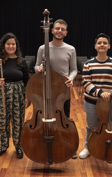 
A imagem mostra os mesmos três jovens músicos no mesmo auditório com fundo de cortina preta e cadeiras dispostas em fileiras. Eles estão alinhados, com seus instrumentos: à esquerda, a jovem com a flauta transversal; ao centro, o rapaz com o contrabaixo; e à direita, o jovem com o violoncelo. O enquadramento é mais amplo, capturando o piso de madeira polido e as cadeiras acolchoadas em maior detalhe. Todos continuam sorrindo, destacando o ambiente descontraído e amigável.