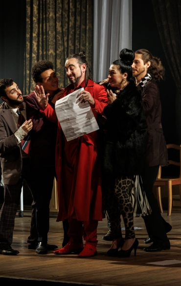 A imagem retrata uma cena de teatro em um palco bem iluminado. No centro, há um homem vestido com um longo casaco vermelho, segurando um papel enquanto faz uma expressão enfática. Ele está cercado por outros atores em trajes formais e elegantes, todos interagindo de maneira dramática.

À esquerda, uma mulher idosa, usando um chapéu grande e preto e apoiada em uma bengala, observa a cena. Ao lado dela, há um homem com barba e terno xadrez olhando intensamente para o homem de vermelho. Um outro homem, de pele mais escura, veste um blazer e sorri enquanto se inclina para o papel.

À direita, uma mulher com cabelo preso em um coque alto e roupa com estampa de oncinha interage com o grupo, enquanto outra, com um vestido roxo brilhante e um cinto dourado, parece intrigada e observa de perto.

Ao fundo, a decoração do palco inclui uma cama hospitalar à esquerda, com lençóis brancos e uma mesa com um castiçal dourado. As cortinas são escuras e pesadas, com uma janela sugerindo um cenário urbano noturno. O chão do palco é de madeira, e algumas folhas de papel estão espalhadas.