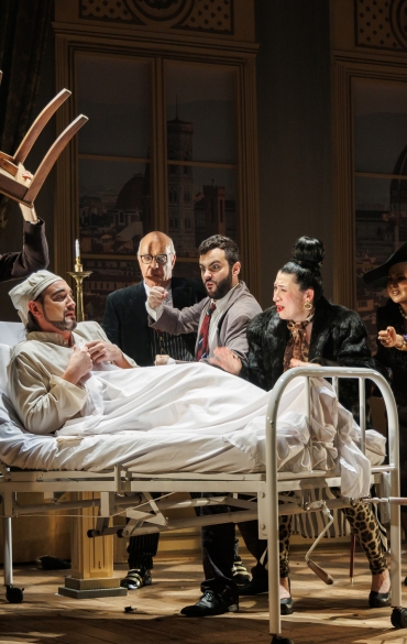 A imagem retrata uma cena de teatro com um grupo de atores em torno de uma cama hospitalar no palco. No centro, um homem está deitado na cama, usando roupas brancas e uma touca, com expressão preocupada, segurando o lençol. Ele é o foco de atenção dos outros personagens.

À esquerda, um homem com cabelos longos, vestindo um casaco escuro com detalhes xadrez, levanta uma cadeira acima da cabeça, aparentando estar em um momento de tensão ou fúria. Ao lado dele, um homem mais velho, calvo e de óculos, observa a cena com um semblante sério.

À direita do homem na cama, um ator de barba, usando um terno, gesticula energicamente, enquanto uma mulher com coque alto e casaco de pele parece angustiada. Atrás dela, outra mulher, de chapéu preto e roupa elegante, esboça um sorriso discreto. Mais à direita, uma mulher de vestido roxo com cinto dourado estende a mão, aparentando preocupação.

O cenário ao fundo é composto por cortinas pesadas e duas grandes janelas, mostrando uma vista de uma cidade histórica com cúpulas e torres. O palco de madeira reforça o ambiente clássico da peça. A iluminação quente destaca o dinamismo e a intensidade da cena teatral.