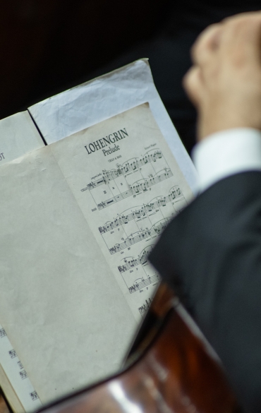 A imagem mostra um músico segurando seu instrumento, provavelmente um violoncelo ou contrabaixo, enquanto lê uma partitura. A página aberta exibe duas obras distintas: "Im Abendrot", uma peça associada a Richard Strauss, e o prelúdio de "Lohengrin", de Richard Wagner. O músico, de terno escuro, está sentado, e o foco da imagem está na partitura e na posição de suas mãos.

O cenário indica um ambiente orquestral, com cadeiras e outros instrumentos parcialmente visíveis. A iluminação parece natural, destacando os detalhes da partitura e criando um clima de concentração e serenidade, típico de momentos de ensaio ou apresentação clássica.