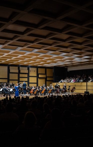 A imagem retrata uma apresentação de orquestra em um auditório. No palco, há diversos músicos tocando instrumentos como violinos, violoncelos, contrabaixos, além de outros instrumentos de uma orquestra sinfônica. Ao centro, uma mulher em um vestido azul longo está em evidência, provavelmente uma solista ou cantora. Ao lado dela, um maestro conduz os músicos.

O ambiente é amplo, com um teto com estrutura geométrica em formato de painéis quadrados. As paredes do fundo e laterais são revestidas com painéis de madeira e detalhes escuros, conferindo uma estética acústica e sofisticada ao local. A plateia está presente em grande número, com muitas pessoas sentadas no nível inferior e outras no balcão superior, assistindo à apresentação. A iluminação é focada no palco, enquanto o público permanece em uma área de sombra. A atmosfera é formal, típica de concertos de música clássica.