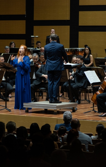 A imagem mostra um close de uma apresentação musical em uma sala de concerto. No centro, uma cantora vestida com um longo vestido azul, de cabelo ruivo, está em plena performance. Ela expressa emoção ao cantar, com gestos amplos e expressivos. Ao seu lado direito está o maestro, vestindo um terno azul escuro, captado de costas enquanto conduz a orquestra com uma batuta levantada.

Ao fundo, membros da orquestra estão concentrados em seus instrumentos, que incluem violinos, clarinetes e trombones. As partituras estão organizadas em estantes diante dos músicos, e o ambiente é sofisticado, com painéis de madeira no fundo, projetados para realçar a acústica. A cena transmite a intensidade e a grandiosidade de uma apresentação clássica.