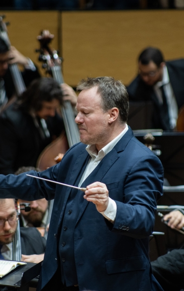 Um maestro está conduzindo uma orquestra em uma sala de concertos. Ele está no centro da imagem, vestindo um terno azul escuro e segurando uma batuta com as mãos em movimento. À sua frente, sobre um púlpito, está uma partitura aberta. Ao fundo, músicos tocam violoncelos e contrabaixos, concentrados em seus instrumentos. O cenário apresenta painéis de madeira clara, e os músicos estão vestidos formalmente, com trajes pretos e gravatas. A expressão do maestro transmite intensidade e foco.