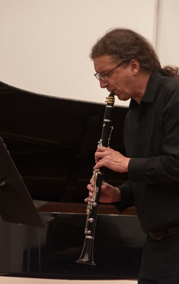 Esta imagem captura um clarinetista em um momento solo durante uma apresentação. Ele está profundamente concentrado, com expressão de dedicação à música. Ao fundo, é possível ver parte de um piano de cauda, que sugere que a performance ocorre em um ambiente de recital ou concerto de câmara.

O clarinetista veste roupas formais, indicando a natureza clássica e refinada do evento. A iluminação quente do ambiente realça a cena, criando uma atmosfera acolhedora e elegante, típica de apresentações de música clássica. Essa proximidade com o público valoriza a interação emocional entre o artista e a audiência, enriquecendo a experiência musical.