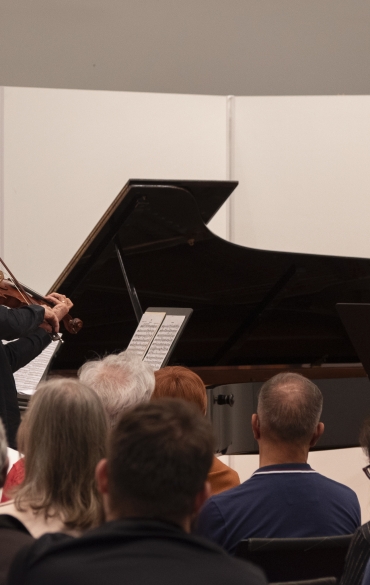 A imagem retrata um recital de música de câmara, com três músicos em destaque: um pianista, um violinista e um clarinetista. Eles estão executando em um espaço intimista, com o público visivelmente próximo ao palco, proporcionando uma atmosfera acolhedora e envolvente.

O pianista está ao fundo, posicionado ao piano de cauda, enquanto o violinista e o clarinetista estão na frente, ambos tocando de forma concentrada. As partituras estão organizadas em estantes, indicando uma apresentação formal e bem estruturada. O público, composto por uma variedade de pessoas, está atento, reforçando a conexão direta entre os músicos e a plateia.

Esse tipo de apresentação geralmente destaca a interação entre os instrumentos e a expressividade musical, sendo uma experiência rica tanto para os artistas quanto para os ouvintes.