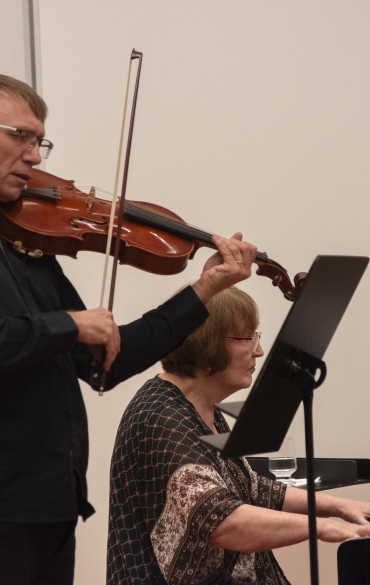 Esta imagem mostra dois músicos em uma apresentação ao vivo. À esquerda, o violinista está concentrado em sua performance, com o arco em movimento sobre as cordas do instrumento, destacando sua habilidade técnica e emoção na execução. À direita, a pianista acompanha no piano de cauda, tocando com expressão e dedicação.

A interação entre os dois músicos reflete a sincronia e a harmonia de um recital de música de câmara. A presença de partituras em suportes evidencia a seriedade da performance e o repertório escolhido. A iluminação suave e o fundo neutro mantêm o foco nos artistas e nos instrumentos, contribuindo para a atmosfera intimista e clássica da apresentação.