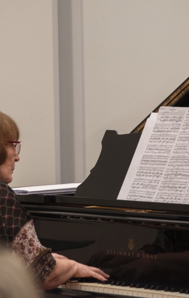 
A imagem mostra uma mulher tocando piano em um ambiente interno. Ela está sentada em frente a um piano de cauda preto da marca "Steinway & Sons". A mulher tem cabelos curtos castanhos e usa óculos vermelhos, além de uma blusa estampada em tons escuros com detalhes geométricos. Sobre o piano, há partituras abertas com várias linhas de notas musicais. O fundo é neutro, com uma parede clara. Na parte inferior da imagem, desfocada, aparece a cabeça de uma pessoa na plateia. O ambiente sugere um recital ou apresentação formal.