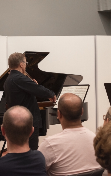 A imagem retrata um recital de música de câmara em um ambiente fechado e iluminado. Em destaque estão três músicos: uma pianista sentada à esquerda, de perfil, tocando um piano de cauda; um violinista de pé no centro, tocando seu instrumento e voltado para a partitura; e um clarinetista à direita, também de pé, tocando enquanto lê uma partitura apoiada em um suporte. Eles vestem roupas escuras, e o fundo do palco é composto por painéis brancos simples. No primeiro plano, vemos a plateia sentada, composta por pessoas de várias idades, que assistem ao recital atentamente. A atmosfera é intimista e cultural.