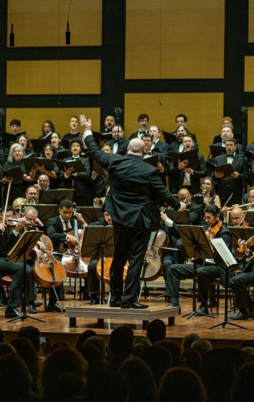 A imagem mostra a Orquestra Sinfônica de Porto Alegre (OSPA) acompanhada de um coro em uma apresentação ao vivo, sob a regência de um maestro. A cena ocorre em um auditório, com a orquestra posicionada na frente e o coro ao fundo. O público pode ser visto na parte inferior, assistindo à performance. A composição transmite um momento de intensidade musical, com os músicos e coristas completamente imersos em sua execução.