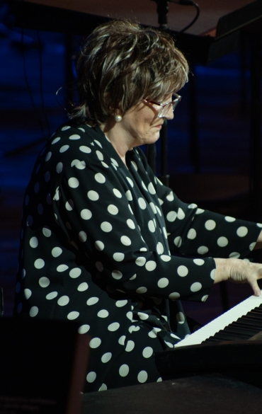 A imagem retrata uma mulher tocando um teclado eletrônico em um ambiente com iluminação teatral. Ela usa óculos de armação clara, brincos de pérola e está vestida com uma blusa preta estampada com bolinhas brancas de tamanho médio. Seus cabelos são curtos e castanhos, com um leve reflexo iluminado pela luz direcionada.

O teclado, identificado como um modelo Roland RD-700, está posicionado sobre um suporte e possui partituras colocadas em um suporte acoplado ao instrumento. Na frente do teclado, há estantes com partituras que exibem o logotipo da Orquestra Sinfônica de Porto Alegre (OSPA).

O fundo do ambiente é escuro, com uma tonalidade azul que sugere iluminação cênica, criando um contraste com a luz mais intensa que destaca a mulher e o teclado. O cenário sugere um contexto de ensaio ou apresentação musical, com foco na expressão artística e no ambiente formal de um concerto.