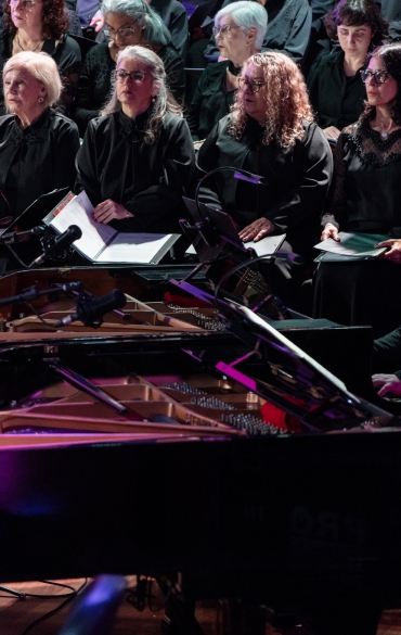 
A imagem captura uma apresentação de música clássica em um ambiente sofisticado, provavelmente em uma sala de concerto ou teatro. Ao fundo, um coro misto composto majoritariamente por mulheres está posicionado em uma arquibancada, todos vestidos em roupas formais predominantemente pretas. As cantoras apresentam expressões concentradas, algumas segurando partituras abertas em suas mãos, sugerindo que estão prontas para cantar ou seguindo a execução da música. A iluminação é suave e elegante, com reflexos de luz roxa que criam uma atmosfera artística e contemplativa.

Na parte da frente, dois pianos de cauda ocupam o primeiro plano da imagem. Os pianos estão posicionados de forma que suas tampas abertas revelam as cordas e os martelos internos, sugerindo que os instrumentos estão sendo utilizados ativamente. Dois pianistas estão sentados frente a frente. O pianista à esquerda veste terno escuro e óculos, com uma expressão de foco absoluto em sua performance. O pianista à direita também está elegantemente vestido com gravata, exibindo um ar concentrado enquanto toca.

A cena é imersiva e carrega um senso de grandiosidade, destacando o esforço colaborativo entre os pianistas e o coro. A disposição dos músicos, a iluminação e o silêncio implícito do público conferem uma sensação de solenidade e beleza típica de concertos de música clássica. A combinação de coral e instrumentos evoca a ideia de uma obra de grande impacto, como uma cantata ou oratória, com provável relevância emocional e cultural.