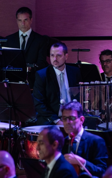 A imagem apresenta uma seção de uma orquestra durante uma performance. No plano de fundo, estão os músicos da percussão, todos vestidos formalmente de terno escuro e gravata. Entre os instrumentos visíveis estão tímpanos, um grande gong e outros instrumentos de percussão. Os percussionistas estão atentos, com expressões sérias e concentradas, como se aguardassem suas entradas na música.

Mais à frente, é possível observar músicos da seção de cordas, como violinistas e violistas, também vestidos formalmente. Alguns estão tocando seus instrumentos, enquanto outros parecem estar aguardando o próximo momento de participação. Os músicos em primeiro plano mostram expressões focadas, enfatizando o clima de precisão e profissionalismo característico de uma orquestra sinfônica.

A iluminação destaca detalhes metálicos dos instrumentos, com tons de luz roxa e quente, criando um contraste com o ambiente predominantemente escuro e reforçando a sofisticação da cena. A disposição dos músicos sugere uma apresentação orquestrada cuidadosamente, possivelmente uma obra grandiosa que exige a interação de várias seções da orquestra, incluindo cordas e percussão.