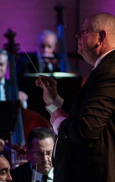 
Esta imagem captura um momento expressivo de um maestro conduzindo uma orquestra em uma apresentação ao vivo. O maestro, em destaque, está concentrado em sua performance, com a batuta levantada, transmitindo sua interpretação musical aos músicos. Atrás dele, os instrumentistas de cordas, incluindo contrabaixos e violoncelos, seguem atentamente a direção, contribuindo para a harmonia da execução.

O uso de iluminação roxa e azul no fundo cria uma atmosfera sofisticada e dramática, típica de apresentações de alto nível artístico. A intensidade e a emoção na expressão do maestro e dos músicos refletem a dedicação e o envolvimento com a música, proporcionando um espetáculo impactante para o público.