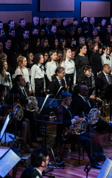 A imagem mostra uma apresentação musical em uma sala de concerto. No primeiro plano, há integrantes de uma orquestra sinfônica sentados em cadeiras com seus instrumentos, como trompas, clarinetes, e instrumentos de corda como violinos e violoncelos. À frente da orquestra, aparecem tambores e outros instrumentos de percussão.

Ao fundo, há um grande coral formado por várias fileiras de cantores. As primeiras fileiras são compostas por jovens vestindo roupas formais (camisas brancas e gravatas ou laços pretos), enquanto as fileiras mais ao fundo apresentam adultos, a maioria vestida de preto.

O ambiente é iluminado por luzes que destacam os músicos e cantores, com painéis acústicos nas paredes. A atmosfera transmite o sentimento de uma apresentação formal e emocionante.