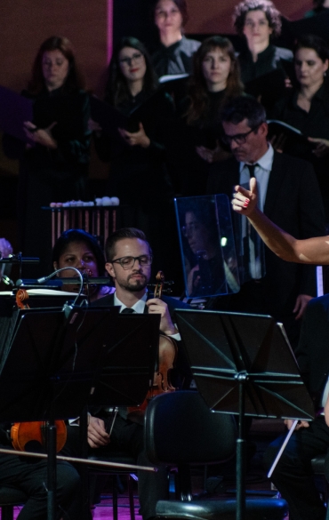 A imagem mostra uma cantora solista em destaque durante uma apresentação musical. Ela está à direita, usando um vestido preto elegante com detalhes brilhantes e está com os braços abertos em um gesto expressivo, transmitindo emoção enquanto canta.

No plano central e à esquerda, músicos da orquestra estão sentados com seus instrumentos, como violinos e violas. Eles estão vestidos formalmente com ternos e gravatas. Ao fundo, é possível ver parte de um coral, composto por cantores vestidos de preto, segurando partituras.

A iluminação destaca a solista e os músicos, criando um ambiente cênico e envolvente, típico de uma performance clássica.