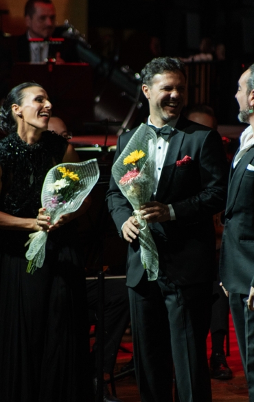 Esta foto mostra um momento de celebração em um concerto, possivelmente no encerramento de uma apresentação. Três artistas estão no centro da imagem, segurando buquês de flores, um gesto tradicional de reconhecimento e agradecimento no meio artístico. Eles sorriem amplamente, demonstrando a alegria e a gratidão pela recepção calorosa do público.

No fundo, a presença da orquestra adiciona profundidade à cena, com músicos e instrumentos compondo um cenário sofisticado. A iluminação quente e os tons avermelhados criam uma atmosfera acolhedora e realçam a energia festiva do momento. A interação dos artistas reflete o espírito colaborativo e a emoção compartilhada que marcam apresentações musicais de grande impacto.