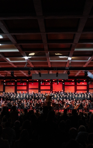 A imagem retrata uma vista ampla de uma sala de concerto durante uma apresentação musical. No palco, uma orquestra sinfônica está posicionada à frente, com músicos tocando seus instrumentos de cordas, sopros e percussão. Atrás da orquestra, há um grande coral, formado por várias fileiras de cantores vestidos de preto.

A iluminação cênica é predominante em tons de vermelho e dourado, destacando o fundo do palco e criando um ambiente dramático. O teto apresenta uma estrutura quadriculada, com refletores apontados para diferentes áreas do palco.

O público aparece em silhueta nas cadeiras da plateia, evidenciando a concentração e o engajamento dos espectadores com a performance. No centro do palco, o maestro está de pé, conduzindo o espetáculo com movimentos amplos.