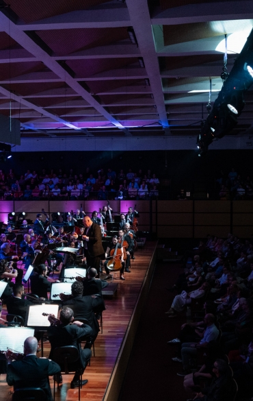 
Essa foto captura a grandiosidade de um concerto orquestral em um auditório. O enquadramento destaca a interação entre a orquestra e o coral, com o maestro em posição de liderança no centro do palco. A plateia, composta por um grande número de espectadores, confere à cena uma atmosfera de envolvimento e prestígio.

O design arquitetônico do auditório, com teto geométrico e iluminação cênica, complementa a estética visual, enquanto os tons arroxeados e azulados da luz criam uma atmosfera dramática e sofisticada. É uma composição que transmite a energia de uma performance ao vivo, com músicos e espectadores imersos na experiência.