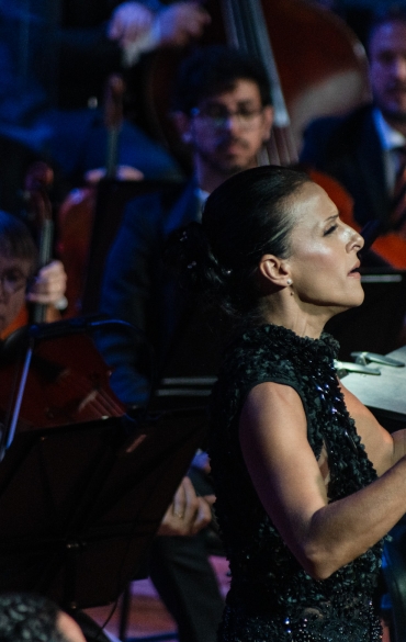 A imagem mostra uma apresentação de uma orquestra sinfônica em um momento de grande expressividade. No centro, uma maestrina conduz com gestos amplos e intensos, usando as mãos para orientar os músicos. Ela está vestida com um vestido preto elegante e brilha sob a iluminação cênica. Ao fundo, vemos músicos de cordas e de contrabaixo, todos vestidos formalmente, acompanhando atentamente a regência. Um dos músicos utiliza máscara facial branca, destacando-se no conjunto. As partituras estão dispostas em suportes à frente dos músicos. O ambiente tem uma iluminação dramática, com tons azulados que acentuam a atmosfera solene e artística do evento.