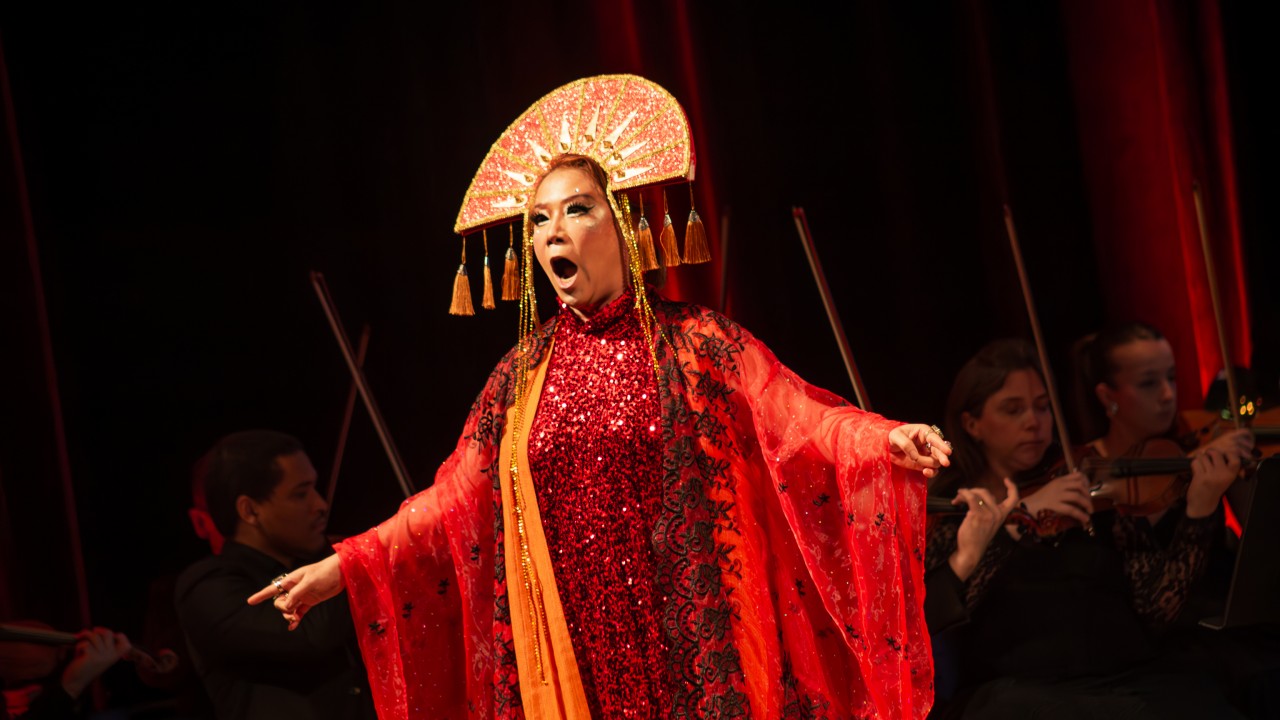 Imagem de uma apresentação de ópera no palco. No centro, uma cantora lírica está em destaque, vestida com um figurino exuberante e marcante. Ela usa um vestido longo e brilhante em tons de vermelho e laranja, coberto por uma capa transparente com detalhes em preto. Na cabeça, ela ostenta um adereço dourado e em formato de leque, com franjas penduradas, remetendo a elementos orientais. Sua expressão facial é intensa, com a boca aberta enquanto canta, e os braços abertos, transmitindo emoção e dramaticidade. Ao fundo, membros de uma orquestra, incluindo violinistas, tocam seus instrumentos, todos vestidos em trajes pretos. O palco é iluminado com luzes vermelhas, reforçando a atmosfera dramática e grandiosa da cena.