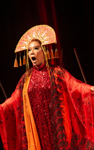 Imagem de uma apresentação de ópera no palco. No centro, uma cantora lírica está em destaque, vestida com um figurino exuberante e marcante. Ela usa um vestido longo e brilhante em tons de vermelho e laranja, coberto por uma capa transparente com detalhes em preto. Na cabeça, ela ostenta um adereço dourado e em formato de leque, com franjas penduradas, remetendo a elementos orientais. Sua expressão facial é intensa, com a boca aberta enquanto canta, e os braços abertos, transmitindo emoção e dramaticidade. Ao fundo, membros de uma orquestra, incluindo violinistas, tocam seus instrumentos, todos vestidos em trajes pretos. O palco é iluminado com luzes vermelhas, reforçando a atmosfera dramática e grandiosa da cena.