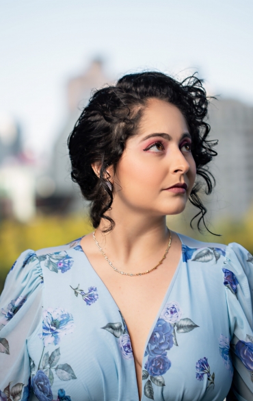 A imagem mostra uma mulher de pele clara e cabelos escuros e cacheados, com um penteado parcialmente preso, posando ao ar livre durante o dia. Ela usa uma maquiagem marcante, com sombra rosa e batom discreto. Seu olhar está direcionado para o lado, transmitindo uma expressão serena e contemplativa. Ela veste um vestido azul claro com estampa floral azul e roxa, de mangas bufantes e decote em "V". Ao fundo, há um ambiente urbano desfocado, com prédios e árvores, sugerindo um cenário de cidade com áreas verdes. A iluminação suave realça seus traços e o tecido do vestido.