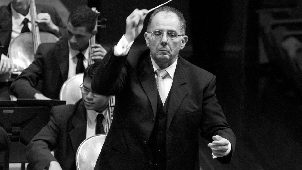 A imagem em preto e branco mostra um maestro em primeiro plano conduzindo uma orquestra. Ele está vestindo um terno escuro com colete e gravata clara. O maestro segura uma batuta com a mão direita levantada, enquanto a mão esquerda está ligeiramente mais baixa, em movimento. Seu rosto expressa concentração e seriedade. Ao fundo, vê-se músicos da orquestra tocando instrumentos de corda, como violoncelos, também vestidos formalmente com ternos escuros e gravatas. A cena acontece em um ambiente de concerto, com iluminação suave focada nos músicos.