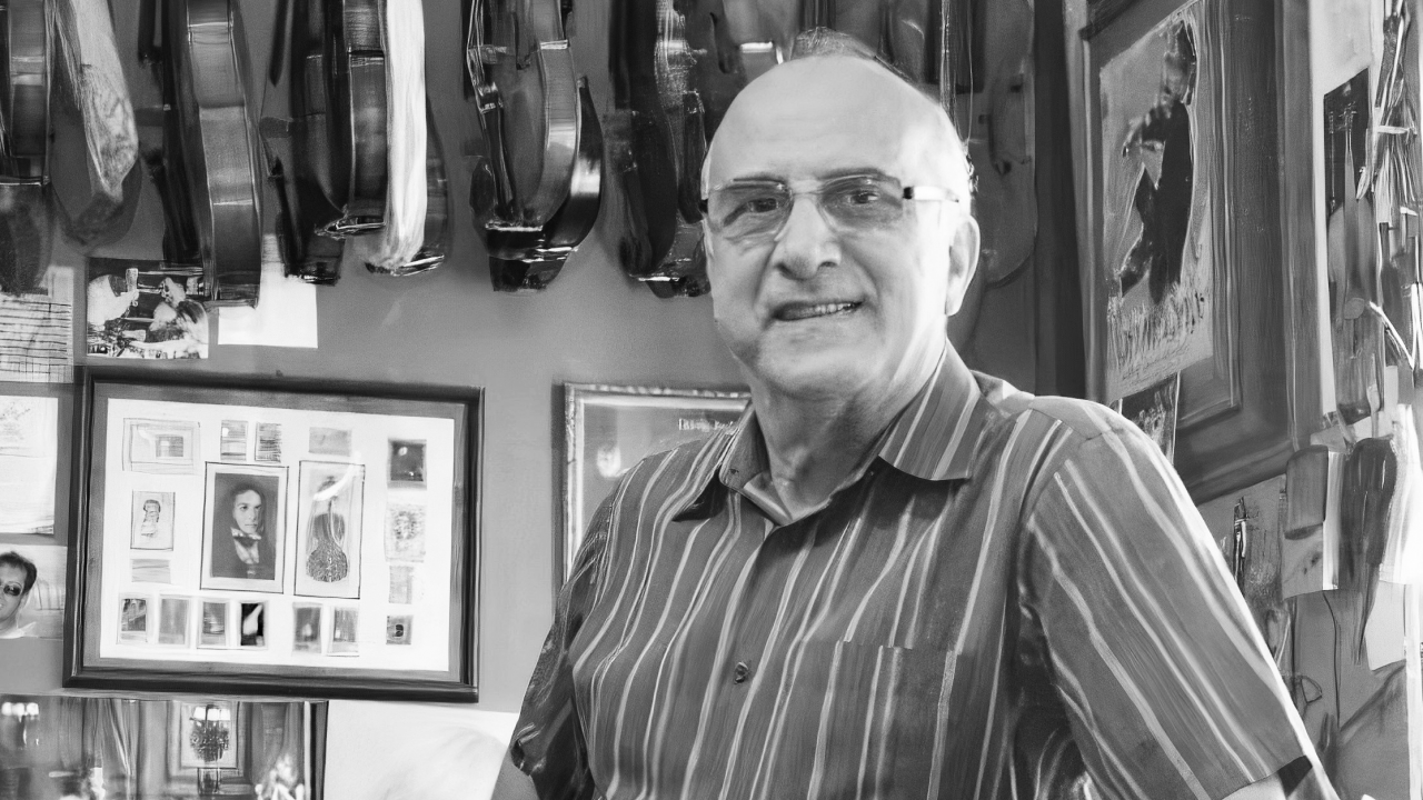 Fotografia em preto e branco de um homem mais velho, calvo e usando óculos, posando sorridente em um ambiente repleto de objetos relacionados à música. Ele veste uma camisa de manga curta listrada e está de pé, levemente inclinado para frente com um dos braços apoiado. Atrás dele, há várias violas e violinos pendurados na parede, além de muitos quadros, fotos e recortes. O espaço parece ser uma oficina ou estúdio de luthieria, com ferramentas e imagens que remetem ao universo musical. A iluminação natural entra pela lateral direita da imagem.