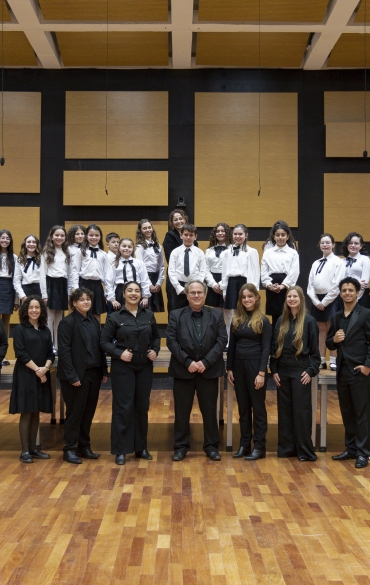 A imagem mostra um grupo grande de pessoas posando para uma foto em um ambiente interno, provavelmente um audit&oacute;rio ou sala de concertos com piso de madeira e paredes com pain&eacute;is ac&uacute;sticos em preto e madeira clara. O grupo est&aacute; dividido em duas fileiras principais.

Na fileira superior, est&atilde;o jovens &ndash; crian&ccedil;as e adolescentes &ndash; uniformizados com camisas brancas, saias ou cal&ccedil;as escuras e gravatas pretas. Eles est&atilde;o dispostos em uma arquibancada de cor clara.

Na fileira inferior, &agrave; frente, est&atilde;o adultos e alguns adolescentes, vestidos predominantemente de preto, com express&otilde;es sorridentes ou neutras. Provavelmente s&atilde;o professores, regentes e membros mais velhos do grupo musical.

O ambiente sugere que o grupo faz parte de um coral infantojuvenil, possivelmente ap&oacute;s uma apresenta&ccedil;&atilde;o ou ensaio.