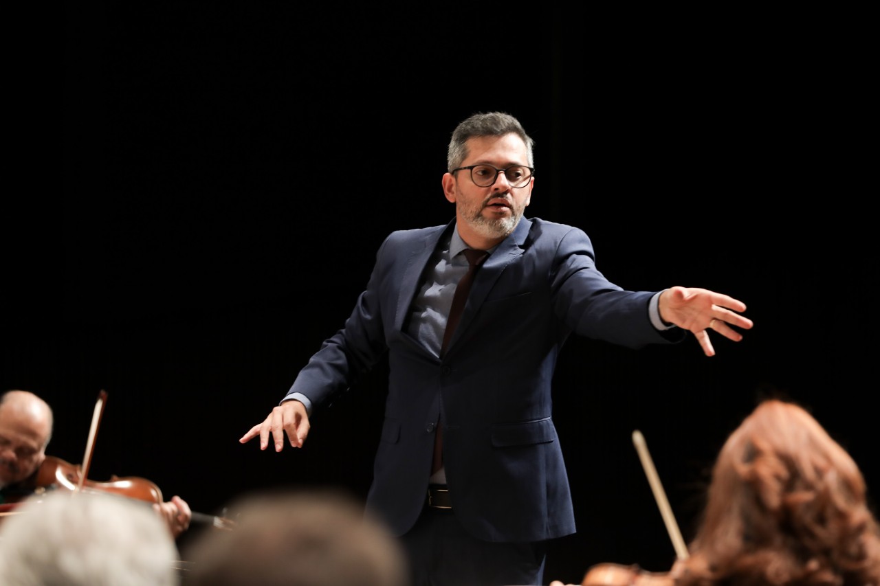 Foto do maestro Rossini Parucci regendo uma orquestra. Ele é um homem de pele clara, cabelos e barba grisalhos, vestindo um terno azul-marinho e óculos de armação escura. Ele está posicionado ao centro, com uma expressão de profunda concentração e os braços estendidos em um gesto de comando direcionado aos músicos. O fundo é escuro, destacando sua figura sob a iluminação do palco, enquanto em primeiro plano aparecem partes de músicos e seus instrumentos, como violinos, levemente desfocados.