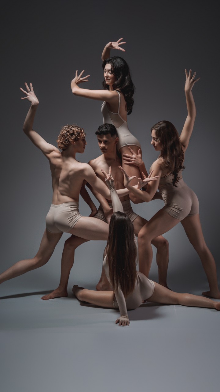 A imagem apresenta um grupo de cinco bailarinos da Cia Carol Dalmolin em uma composi&ccedil;&atilde;o art&iacute;stica sobre um fundo cinza. Os artistas, quatro mulheres e um homem, est&atilde;o agrupados ao centro em diferentes n&iacute;veis &mdash; alguns em p&eacute; e uma bailarina ajoelhada &agrave; frente, formando uma escultura humana harmoniosa. Todos vestem figurinos minimalistas em tons de bege.