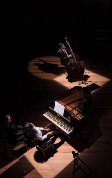 A imagem registra uma vista a&eacute;rea de um quinteto musical em uma apresenta&ccedil;&atilde;o intimista sobre um palco de madeira clara. O ambiente est&aacute; predominantemente &agrave;s escuras, com a ilumina&ccedil;&atilde;o composta por feixes de luz direcionais que isolam cada m&uacute;sico em c&iacute;rculos brilhantes, gerando sombras dram&aacute;ticas e alongadas no ch&atilde;o. Na parte inferior esquerda, duas mulheres ocupam um piano de cauda preto, sendo que uma delas toca o instrumento enquanto a outra observa logo atr&aacute;s. No canto superior esquerdo, um m&uacute;sico toca um contrabaixo ac&uacute;stico, enquanto no lado oposto, ao fundo, um violinista permanece de p&eacute; diante de sua estante de partituras. &Agrave; direita, em primeiro plano, um percussionista aparece de costas, posicionado sobre um tapete escuro com seu kit de bateria. A disposi&ccedil;&atilde;o dos artistas forma um semic&iacute;rculo que, aliado &agrave; presen&ccedil;a discreta de equipamentos de grava&ccedil;&atilde;o como trip&eacute;s e microfones, sugere a produ&ccedil;&atilde;o de um concerto de m&uacute;sica de c&acirc;mara ou jazz em um est&uacute;dio ou audit&oacute;rio.
