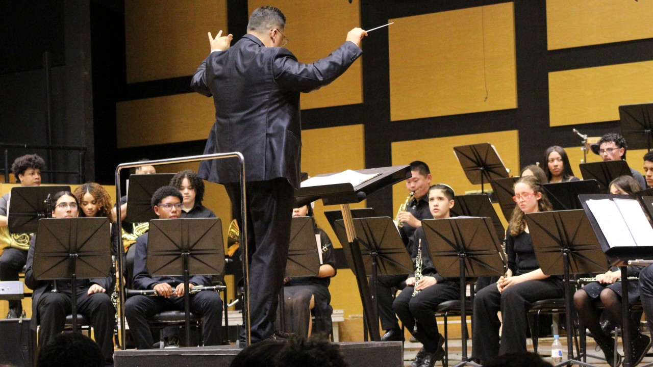 A imagem registra o regente Wilthon Matos de costas para a c&acirc;mera, conduzindo a Orquestra de Sopros da Escola da OSPA sobre um pequeno tablado preto. Ele veste um terno escuro e gesticula com os bra&ccedil;os erguidos, segurando uma batuta na m&atilde;o direita enquanto orienta um grupo de jovens m&uacute;sicos. Os instrumentistas, trajando roupas pretas formais, est&atilde;o organizados em semic&iacute;rculo com seus instrumentos de sopro e partituras, mantendo o olhar atento &agrave; reg&ecirc;ncia. Ao fundo, a parede do palco apresenta um design geom&eacute;trico com pain&eacute;is de madeira clara e divis&oacute;rias pretas, enquanto a base da foto mostra, de forma desfocada, o topo das cabe&ccedil;as de alguns espectadores na plateia.